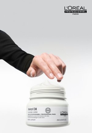 Hand som sträcker sig mot en öppen burk med L'Oréal Professionnel Metal DX professionell hårmask med kräm synlig inuti.