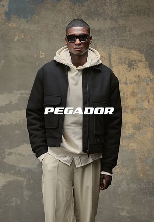 Mann trägt schwarze Jacke über beigem Kapuzenpullover und Hose, mit Sonnenbrille, steht vor einer strukturierten grauen und gelben Wand. Text "PEGADOR" auf der Brust.