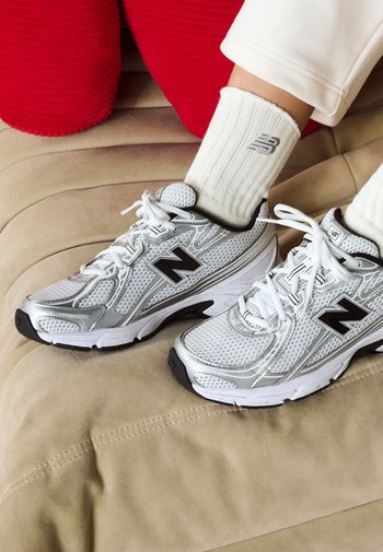 Silberne Sportschuhe mit Netz- und Synthetiküberzügen, mit einem auffälligen schwarzen 'N'-Logo, auf einer strukturierten beige Oberfläche platziert.