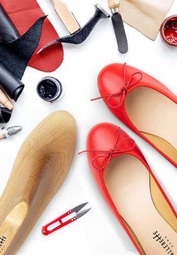 Ballerines en cuir rouge avec un nœud, un moule en bois, et des outils de bricolage incluant des ciseaux et des morceaux de cuir sur un fond blanc.