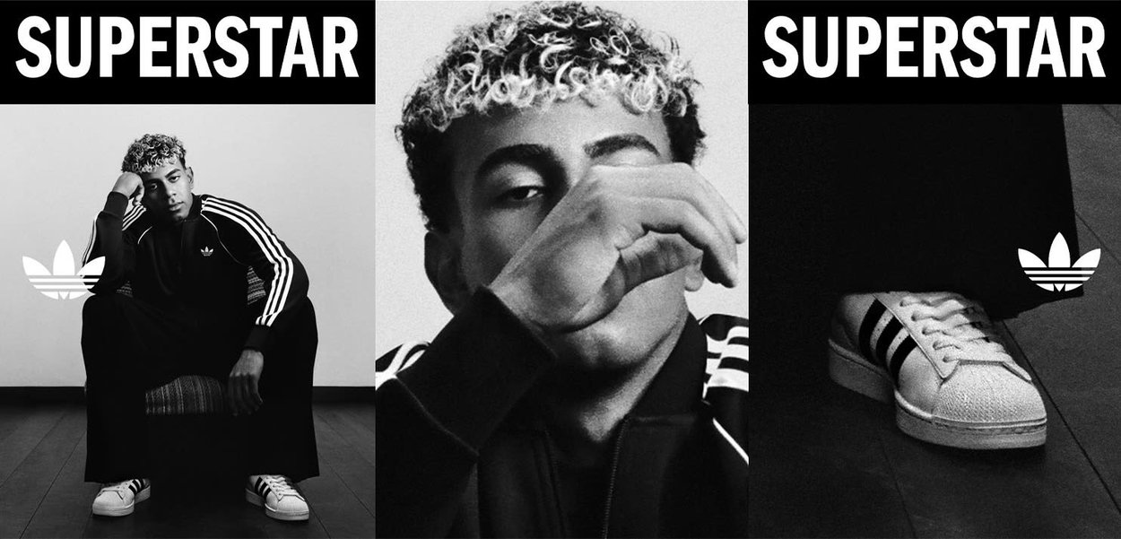 Joven con cabello rizado, vestido con chándal Adidas y zapatillas deportivas, sentado, primer plano del rostro y una sola zapatilla en el suelo con el texto "SUPERSTAR".