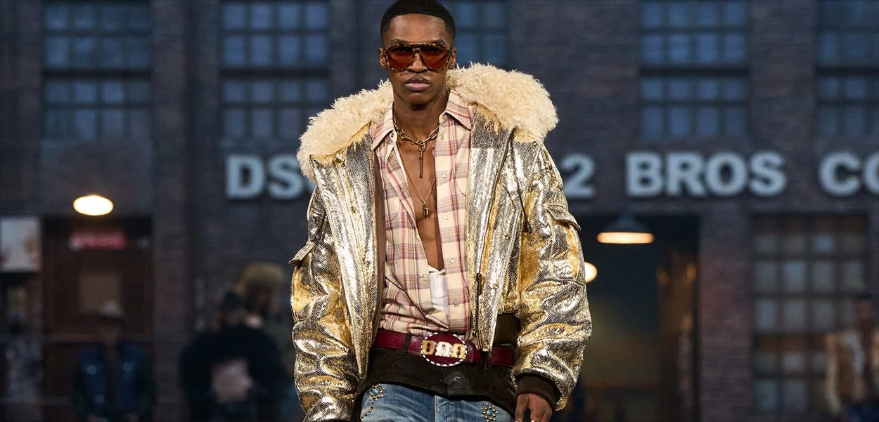 Mann trägt übergroße goldene Metalljacke mit Lammfellkragen, kariertes Hemd, roter Gürtel, Sonnenbrille und Kreuzketten, geht draußen spazieren.