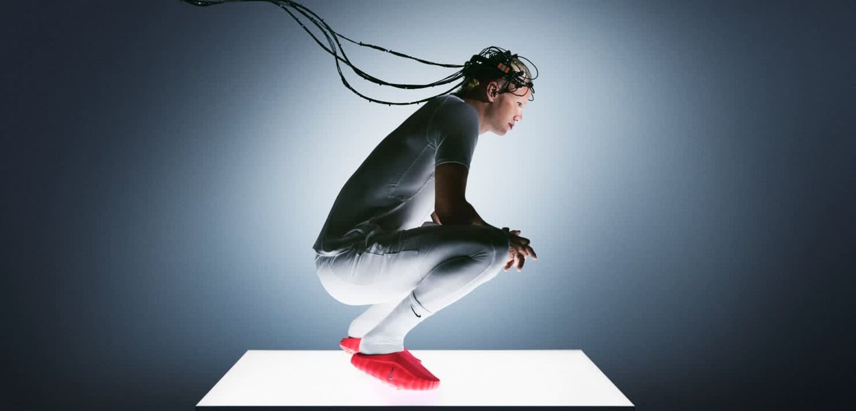 Indumento bianco aderente, calzature rosse e lunghi cavi neri che si estendono dalla testa. Il soggetto è accovacciato su una piattaforma rettangolare illuminata.