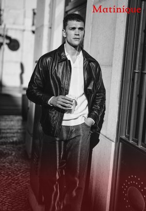 Schwarze Lederjacke über einem weißen Hemd mit Kragen, kombiniert mit grauen Jeans. Die Figur hält eine Tasse und steht in der Nähe einer Türöffnung. Monochromer Ton.