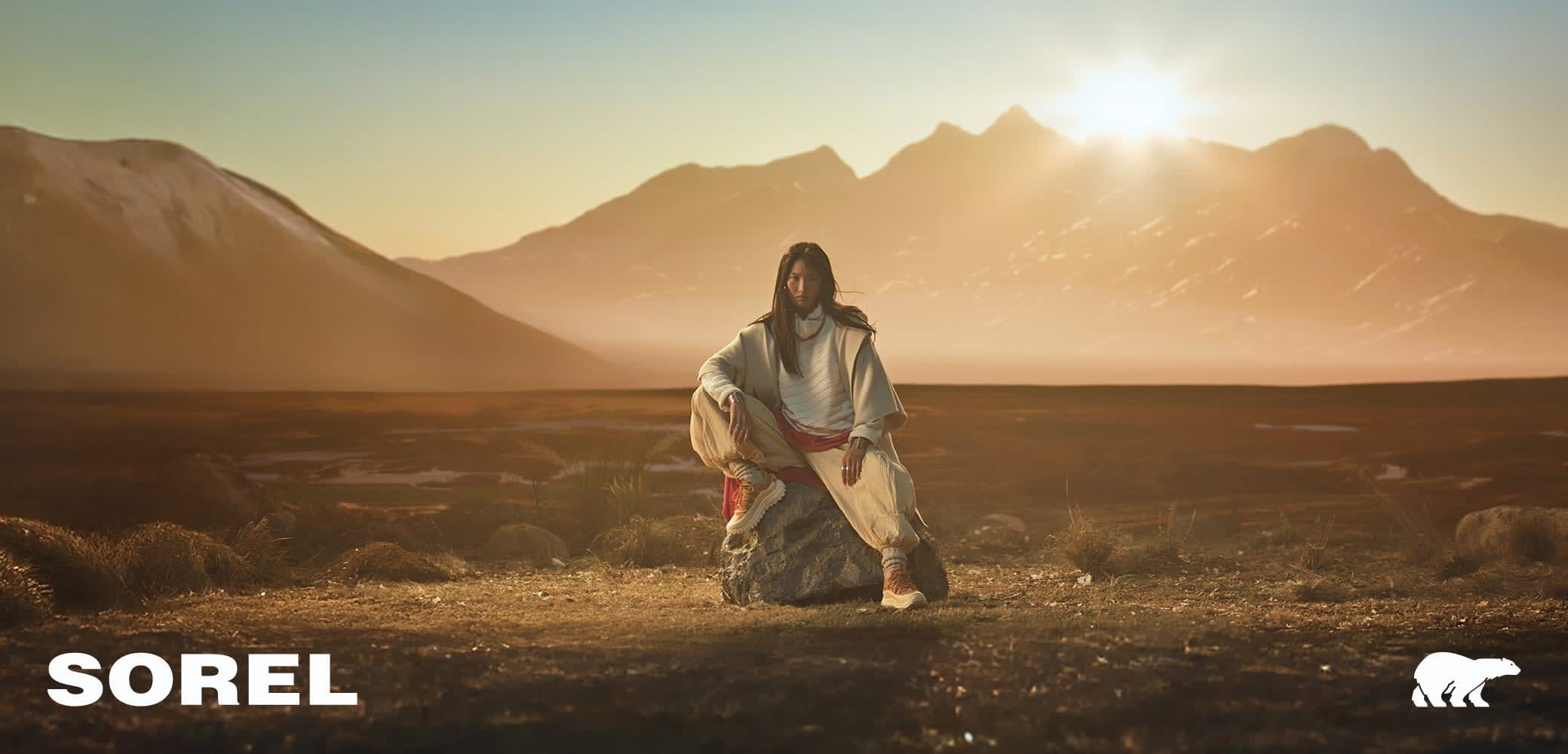 Les chaussures SOREL présentent un design robuste dans des teintes terreuses, avec une semelle en caoutchouc texturé et un dessus protecteur, le tout dans un décor de montagnes ensoleillées.