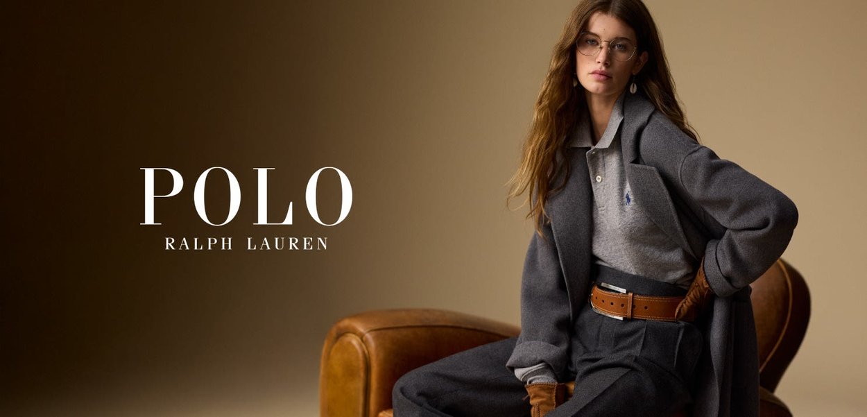 Manteau gris sur une polo gris, ceinture en cuir brun et gants bruns. Le modèle est assis sur une chaise en cuir brun devant un fond neutre.