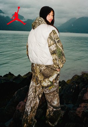 Woman wearing camouflage outfit with white vest stands on rocky shore, facing lake and mountains under cloudy sky, red logo in top left.