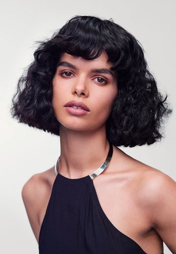 Black curly bob hairstyle, smooth skin, neutral makeup, wearing a sleeveless black top and a silver collar necklace against a plain background.