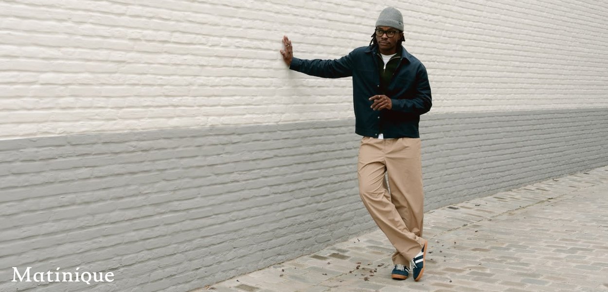 Mann mit grauer Mütze, dunkler Jacke, beigen Hosen und Turnschuhen lehnt an einer zweifarbigen grauen Backsteinmauer auf einem Kopfsteinpflasterweg.