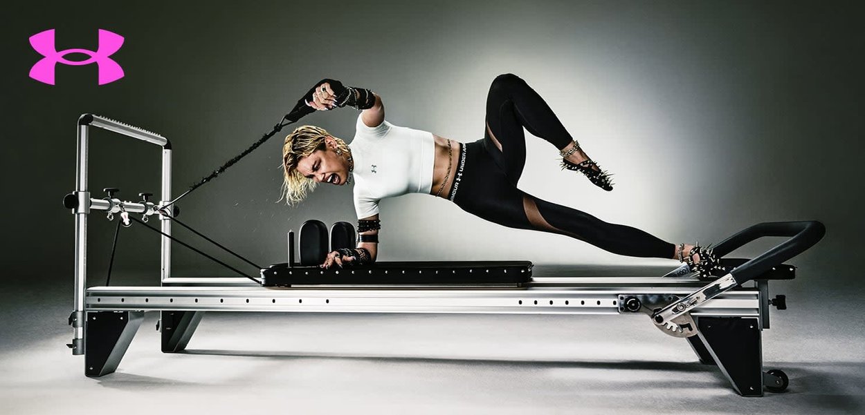 Vrouw in zwarte legging en wit shirt die een reformer Pilates-apparaat gebruikt, waarbij ze weerstandbanden trekt terwijl ze een plankhouding aanhoudt, met zichtbaar Under Armour-logo.