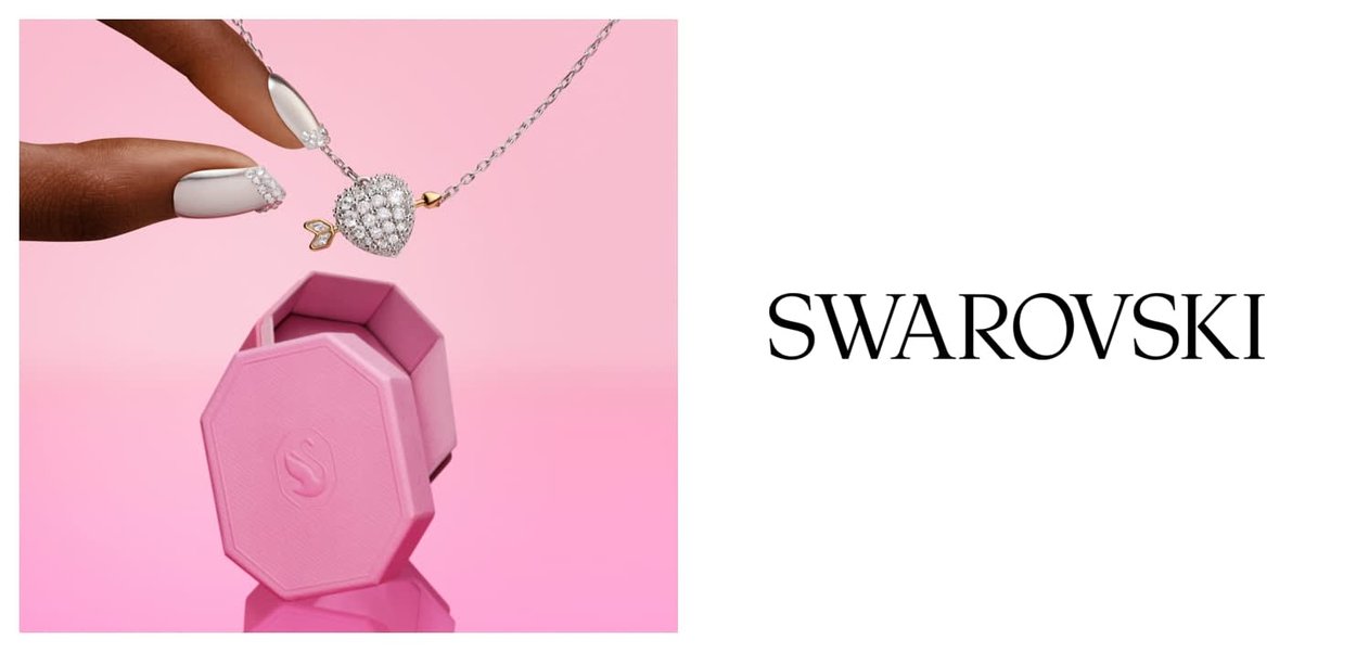 Hand met wit versierde nagels die een hartvormige kristallen hanger vasthoudt boven een roze sieradendoosje, met Swarovski-logo op een witte achtergrond.