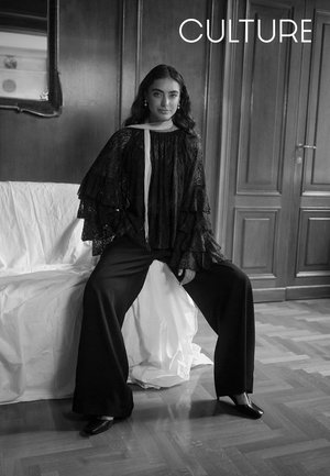 Schwarzes Spitzenoberteil mit geschichteten Rüschen, kombiniert mit schwarzen, weit geschnittenen Hosen. Modell sitzt auf weißem Stoff vor holzfarbenem Hintergrund.