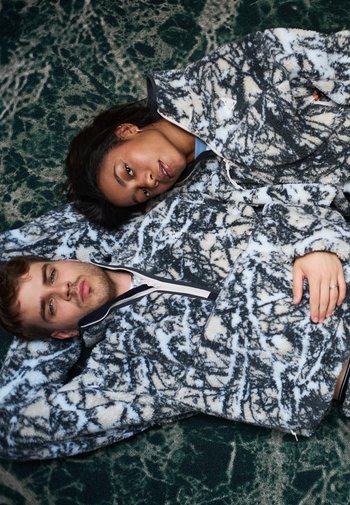 Fleece jassen met een draaikolke patroon in zwart en wit, met een ritssluiting aan de voorkant en hoge kraag, gedragen door twee personen die op een gestructureerd groene ondergrond liggen.