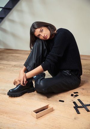 Stivaletti neri in pelle con suole spesse, abbinati a pantaloni neri in pelle e a un maglione a maglia strutturata, posati su un pavimento in legno.