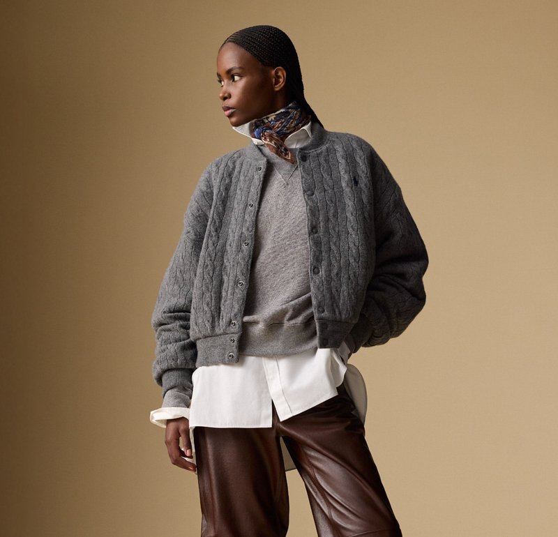 Femme avec des cheveux tressés portant un pull en tricot gris à mailles, un pull gris superposé sur une chemise blanche, un pantalon en cuir marron et une écharpe à motifs.