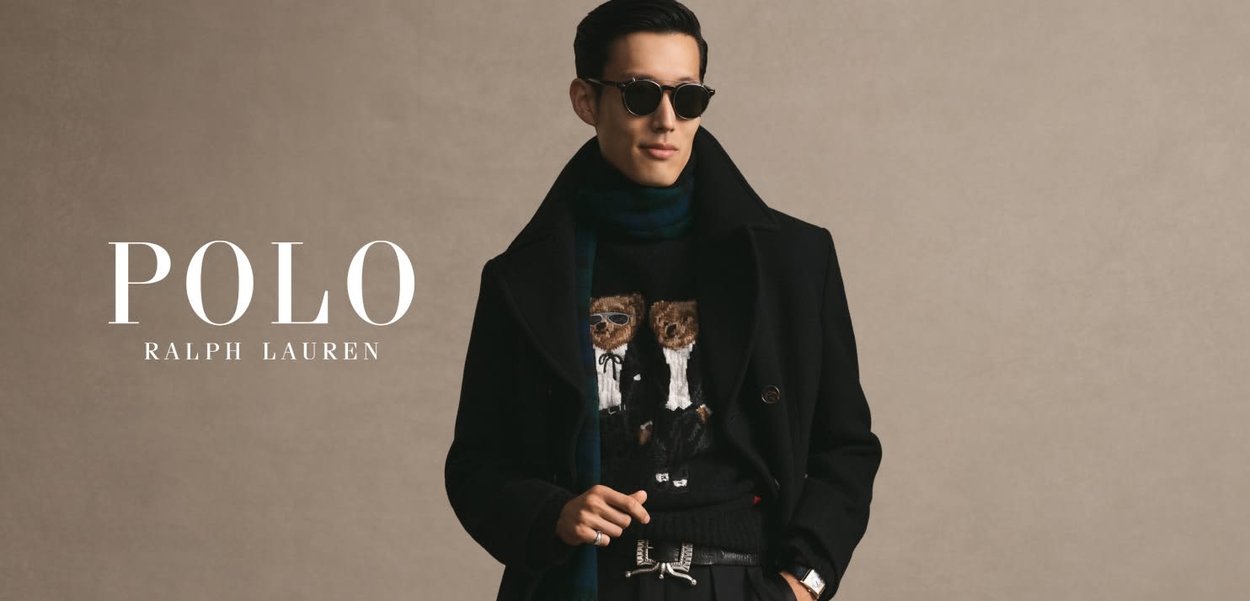 Uomo con cappotto nero, maglione con due orsetti, occhiali da sole, sciarpa e orologio, in piedi davanti a uno sfondo beige uniforme.