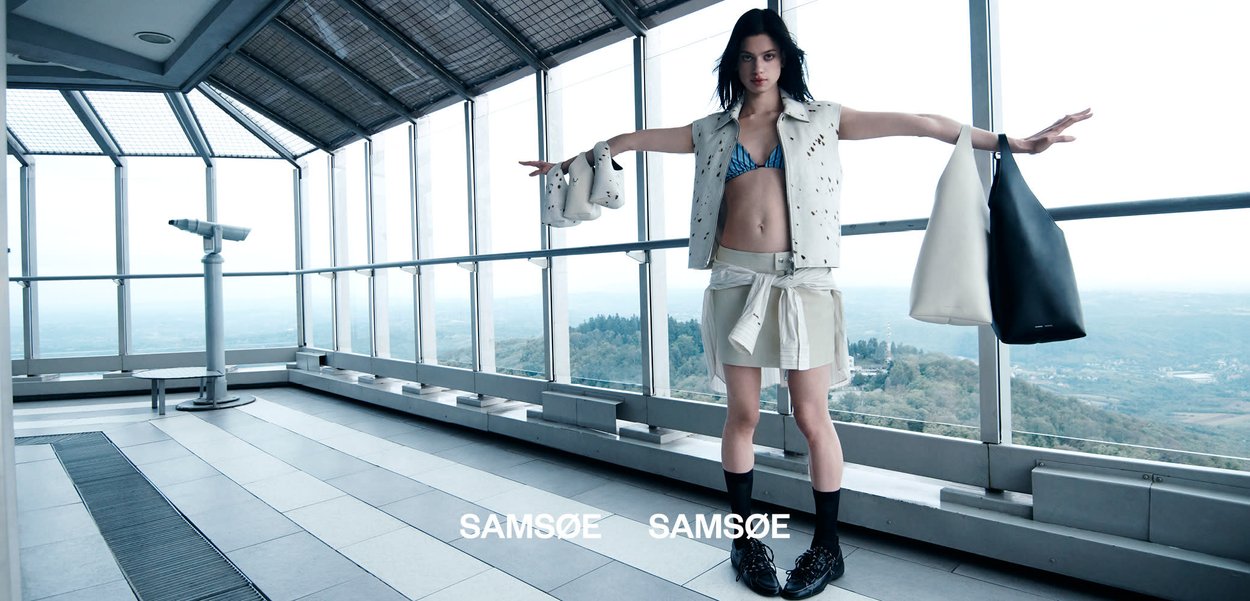 Ung kvinde i moderne påklædning står med armene strakt ud, holder to tasker, på en lys observationsdæk med glasvægge med udsigt over et landskab.