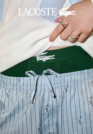 Una mano solleva una camicia bianca per mostrare una fascia verde con il logo del coccodrillo Lacoste bianco sopra pantaloni azzurri a righe con coulisse.