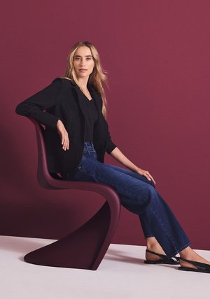 Buet bordeaux stol, tekstureret sort blazer, sort top og flarede blå jeans. Glat gulv og vinrød baggrund fremhæver scenen.