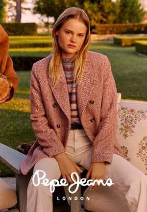 Rosa Fischgräten-Mantel mit doppelter Knopfleiste über einem gestreiften Pullover, kombiniert mit cremefarbenen Hosen. Außenszene mit Grün im Hintergrund.