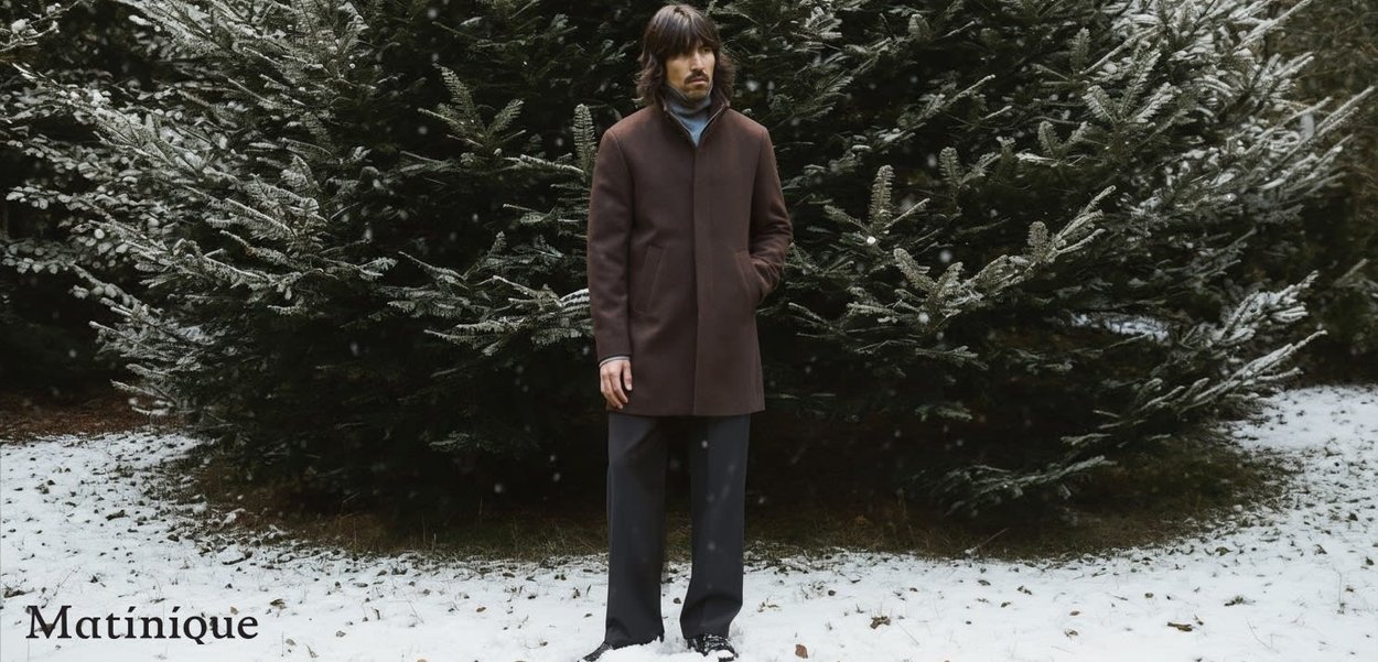 Brown wool blend coat with a high collar and side pockets, worn over a light blue turtleneck, paired with dark trousers and shoes, snowy background.