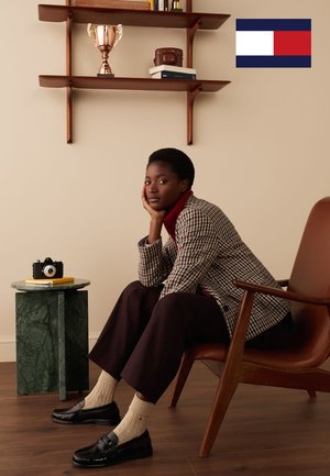 Blazer marron à motif pied-de-poule, col roulé bordeaux, mocassins noirs, chaussettes crème. Table d'appoint en marbre vert avec un appareil photo vintage et un livre. Trophée sur une étagère.