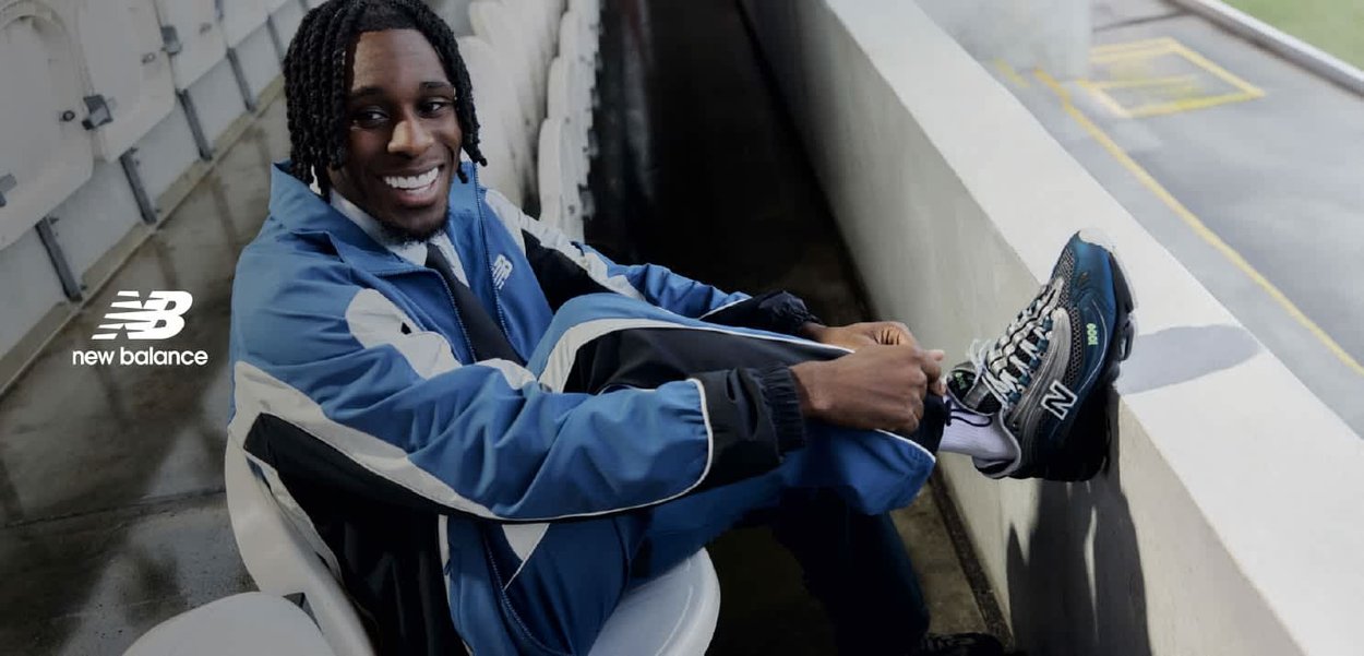 Uomo sorridente in tuta blu e nera che sistema le sneaker blu e grigie New Balance mentre è seduto nei posti all'aperto di uno stadio.