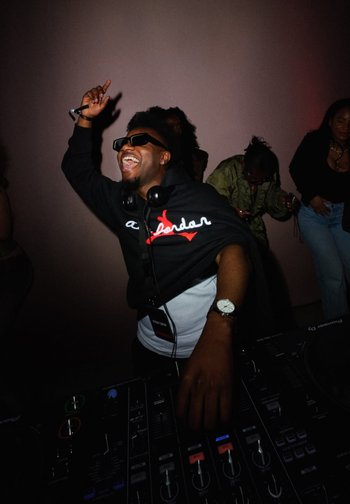 DJ con gafas de sol y auriculares levantando una mano con alegría mientras mezcla música en una consola en una fiesta con poca iluminación y gente bailando.