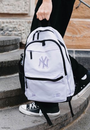 White backpack with black accents and zippers, featuring a purple embroidered logo and two front pockets, held by a person on steps.