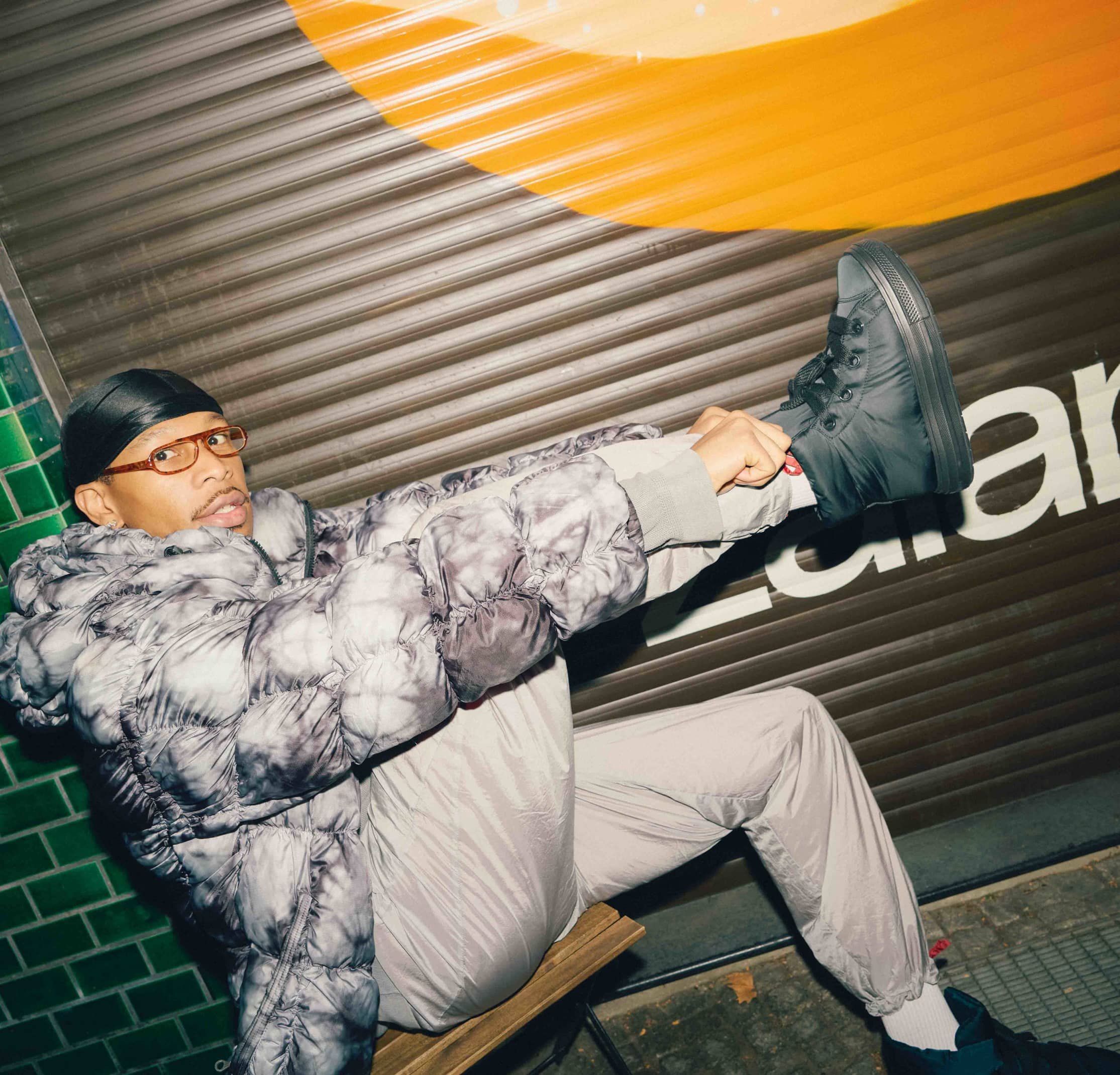 Chaqueta acolchada con un patrón tie-dye, pantalones grises y zapatillas negras altas. El fondo presenta una persiana metálica marrón con acentos naranjas.