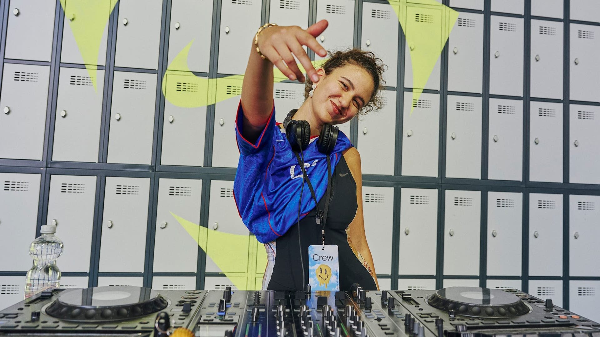Impianto DJ con giradischi neri e mixer su un tavolo. Sullo sfondo ci sono armadietti grigi. Una persona indossa una maglietta blu e cuffie.