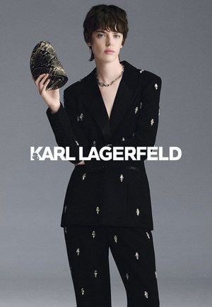 Una modelo con un traje negro adornado sostiene un zapato con estampado de piel de serpiente sobre un fondo gris de estudio. La imagen lleva el nombre de Karl Lagerfeld.