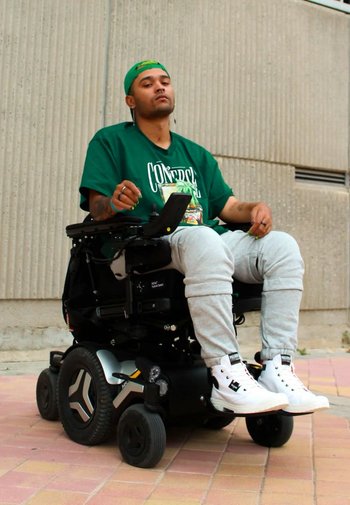 Elektrisk rullstol med svart ram, stora gummihjul och silverdetaljer. Användaren bär en grön t-shirt med grafiskt tryck och ljusgråa sweatpants.