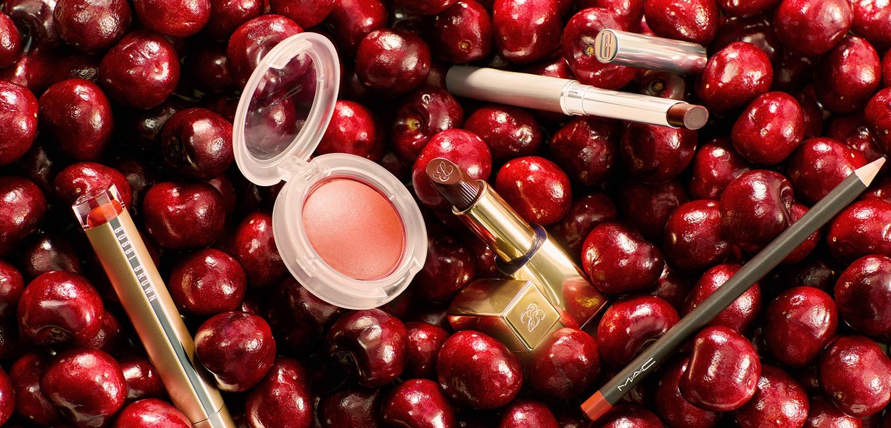Forskellige makeup-produkter, herunder læbepomade, læbestift og blush, spredt ud over en seng af friske, mørkerøde kirsebær.