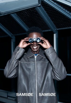 Mann i grå skinnjakke smiler mens han ser gjennom kikkert under en metalltakstruktur om natten.