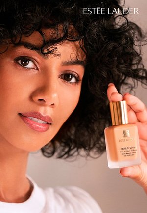 Podkład Double Wear od Estée Lauder w przezroczystej szklanej butelce z złotą zakrętką. Modelka trzyma butelkę i pokazuje gładką teksturę skóry.