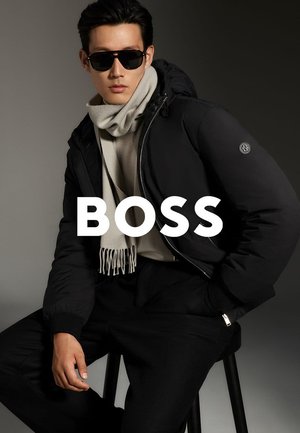 Giacca a vento imbottita nera da uomo con cappuccio, abbinata a una sciarpa grigio chiaro e occhiali da sole. Tessuto liscio, design minimale e dettaglio del logo sulla manica.