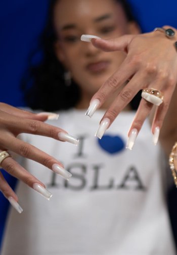 Lange, glanzende amandelvormige nagels met een verloop van wit naar roze. Handen versierd met ringen en een wit grafisch T-shirt tegen een blauwe achtergrond.