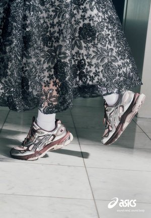Piernas con calcetines blancos que llevan zapatillas Asics combinadas con una falda negra de encaje floral sobre un suelo de baldosas, un pie levantado.