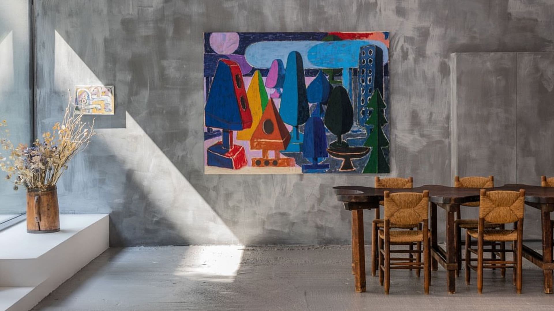 Kleurrijk abstract schilderij met geometrische vormen van bomen; gedeisd grijze muur, houten eettafel met geweven stoelen en een vaas met gedroogde bloemen.