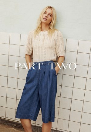 Frau trägt ein cremefarbenes, kurzärmeliges Stricktop und hochtaillierte, lockere blaue Denim-Culotte-Hosen, steht vor einer gefliesten Wand.