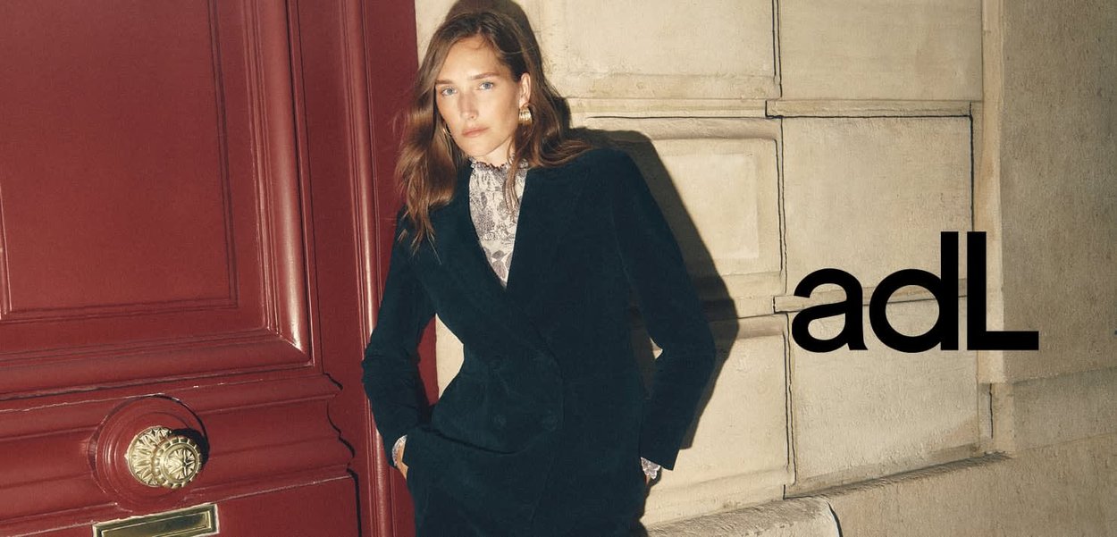 Dunkelgrüner, samtiger, doppelt knöpfbarer Mantel mit seitlichen Taschen und strukturiertem Muster, kombiniert mit einer Spitzbluse, vor einer roten Tür.