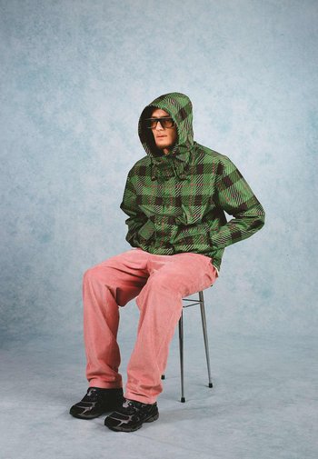 Groen en zwart geruite hoodie met trekkoorden, gecombineerd met roze corduroy broek en zwarte schoenen, persoon zittend op een metalen stoel.