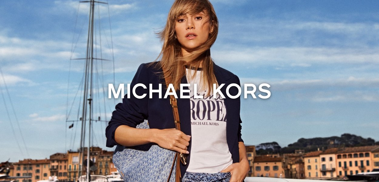 Kvinne iført marineblå blazer og hvit t-skjorte med logo poserer ved marina med yachter og bygninger i bakgrunnen under blå himmel.