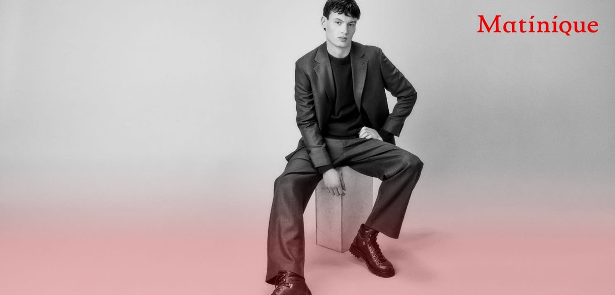 Schwarzer maßgeschneiderter Anzug mit passendem T-Shirt, das elegante Linien aufweist. Das Modell sitzt auf einem hellen Block vor einem sanft verlaufenden Hintergrund.