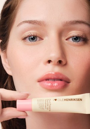 Pfirsichfarbene Tube mit rosa Akzent, beschriftet "Pout Preserve Peptide Lip Treatment", in der Hand einer Person gehalten, nahe dem Gesicht eines Models mit klarer Haut.