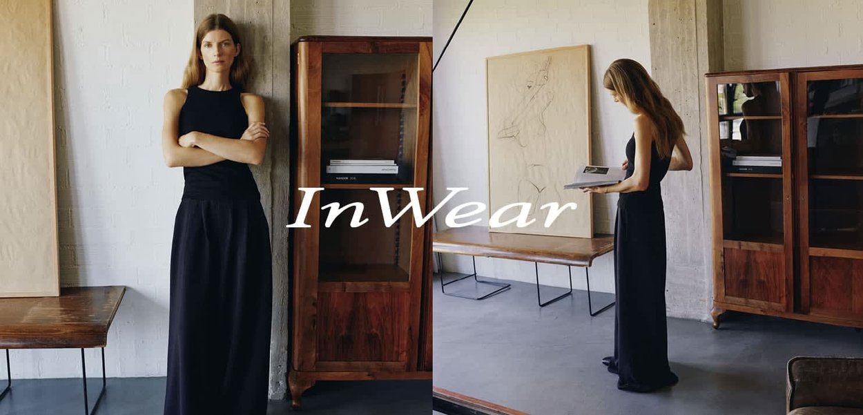 Frau in einem schwarzen ärmellosen Kleid posiert neben einem Holzschrank und liest ein Buch in einem minimalistischen Raum mit hellen Wänden und Holzmöbeln.