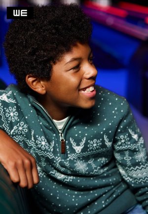 Maglione verde con zip, decorato con un motivo invernale che presenta renne e fiocchi di neve. Il materiale sembra morbido e texturizzato, indossato da un bambino.