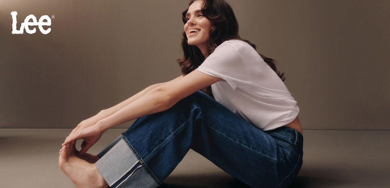 Jeans in denim blu con gambe larghe e risvolti, abbinati a una maglietta bianca. Tessuto seta, posa seduta contro uno sfondo neutro.