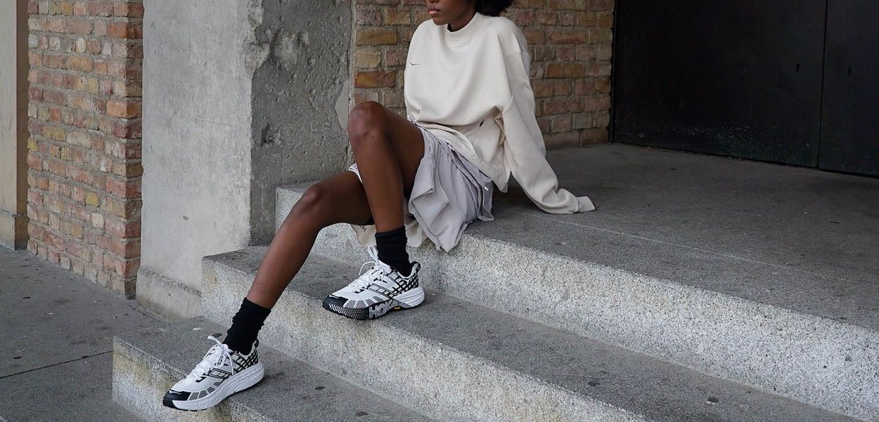 White athletic sneakers with black and geometric accents. Model wears a long beige sweatshirt and layered gray skirt, sitting on stone steps.